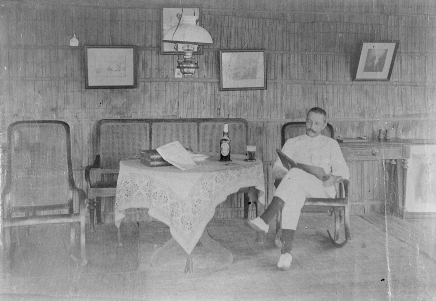 Een lezende man in een woonhuis in de binnenlanden van Borneo met gedekwanden, petroleumlampen en schommelstoelen, 1894 (collectie KITLV 503074)